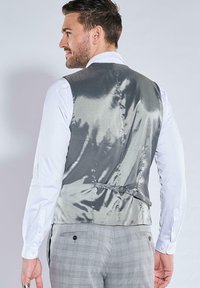 Graue Satinweste mit glatter Textur, mit einer Schnallendetail am Rücken, über einem weißen Hemd getragen. Glatte, maßgeschneiderte Passform.