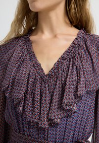 Blauwe blouse met een patroon en een gerimpelde halslijn, met een geometrisch ontwerp in rood en wit. Lichtgewicht stof, zacht gedrapeerd.
