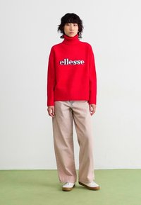 Rode gebreide trui met hoge hals en "ellesse" logo, gecombineerd met lichtbeige wijde broek en witte sneakers op een groene vloer.