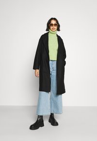 Black coat with a relaxed fit, worn over a green turtleneck, paired with wide-leg light blue jeans and black chunky boots.