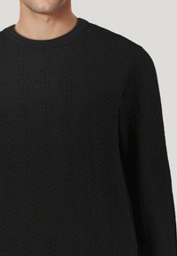 Schwarzer Strickpullover mit einem strukturierten Fischgrätenmuster, Rundhalsausschnitt und langen Ärmeln. Das Material wirkt weich und warm.