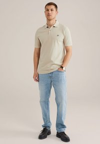 Cămașă polo bej deschis cu mâneci scurte, design minimalist și un mic logo; asortată cu blugi albaștri deschiși și pantofi negri.