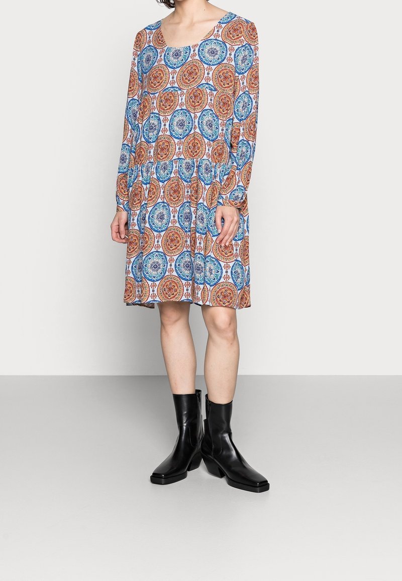 Nainen, joka pukeutunut pitkähihamaiseen mekkoon, jossa on sinisiä ja oransseja pyöreitä kuvioita, ja mustat keskivartalolle ulottuvat nahkasaappaat, seisoo vaalealla lattialla.