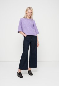 Pull à manches courtes lavande associé à un jean en denim foncé taille haute et jambes larges, porté avec des chaussures noires à talons blocs brillants. Fond blanc épuré.