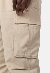 Beige cargo bukser har et teksturert stoff, sidelommer og et merkeetikett på hoften, som viser et praktisk design.
