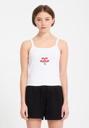 Jeune femme portant un débardeur blanc orné de cœurs rouges et du texte « PAS À TOI », associée à un short noir à cordon, se tenant devant un fond blanc.