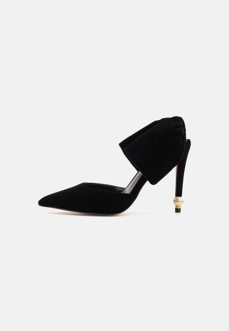 Chaussure à talon haut en velours noir avec bout pointu, large bride chevillée plissée et perle décorative dorée et blanche sur le talon.