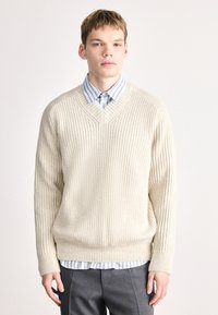 Κρέμα πουλόβερ με πλέξη καλωδίου και V-λαιμόκοψη, σε συνδυασμό με ένα ανοιχτό μπλε με λευκές ρίγες πουκάμισο από κάτω. Γκρι κομμένα παντελόνια ολοκληρώνουν την εμφάνιση.