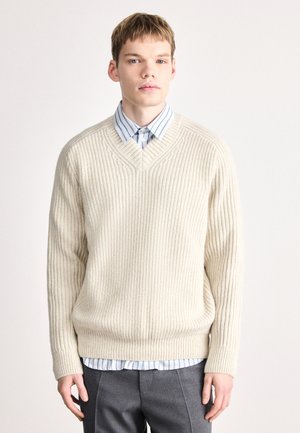 Pull crème en tricot torsadé avec un col en V, associé à une chemise rayée bleu clair et blanche en dessous. Un pantalon gris ajusté complète la tenue.