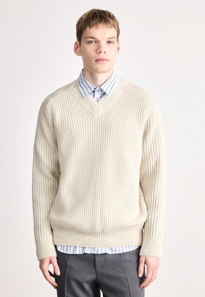 Κρέμα πουλόβερ με πλέξη καλωδίου και V-λαιμόκοψη, σε συνδυασμό με ένα ανοιχτό μπλε με λευκές ρίγες πουκάμισο από κάτω. Γκρι κομμένα παντελόνια ολοκληρώνουν την εμφάνιση.