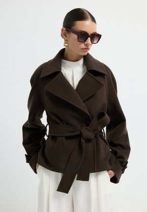 Bruine wollen blend wikkeljas met brede revers, een bijpassende riem en knoopaccenten. Gecombineerd met een witte top en contrasterende brede broek.