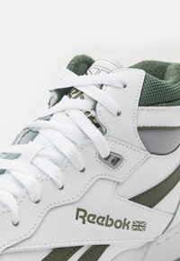 Vita lädersneakers med gröna accenter, texturerat plagg på tungan och vita snören. Reebok-logotypen visas tydligt på sidan.
