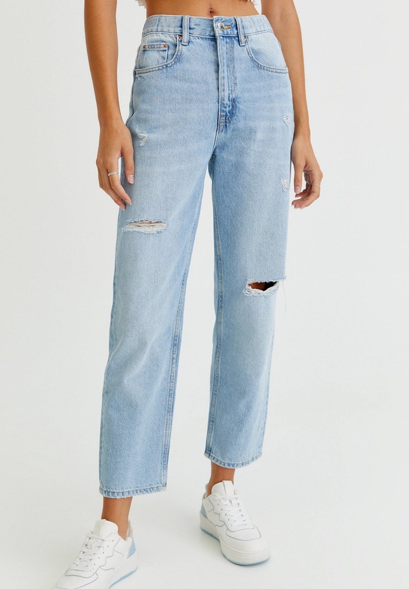 Femme portant un jean déchiré taille haute bleu clair et des baskets compensées blanches, debout devant un fond blanc uni.