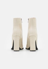 Kremowe botki za kostkę z teksturowanym wzorem krokodyla, wysokim słupkowym obcasem i odsłoniętymi zamkami błyskawicznymi z tyłu.