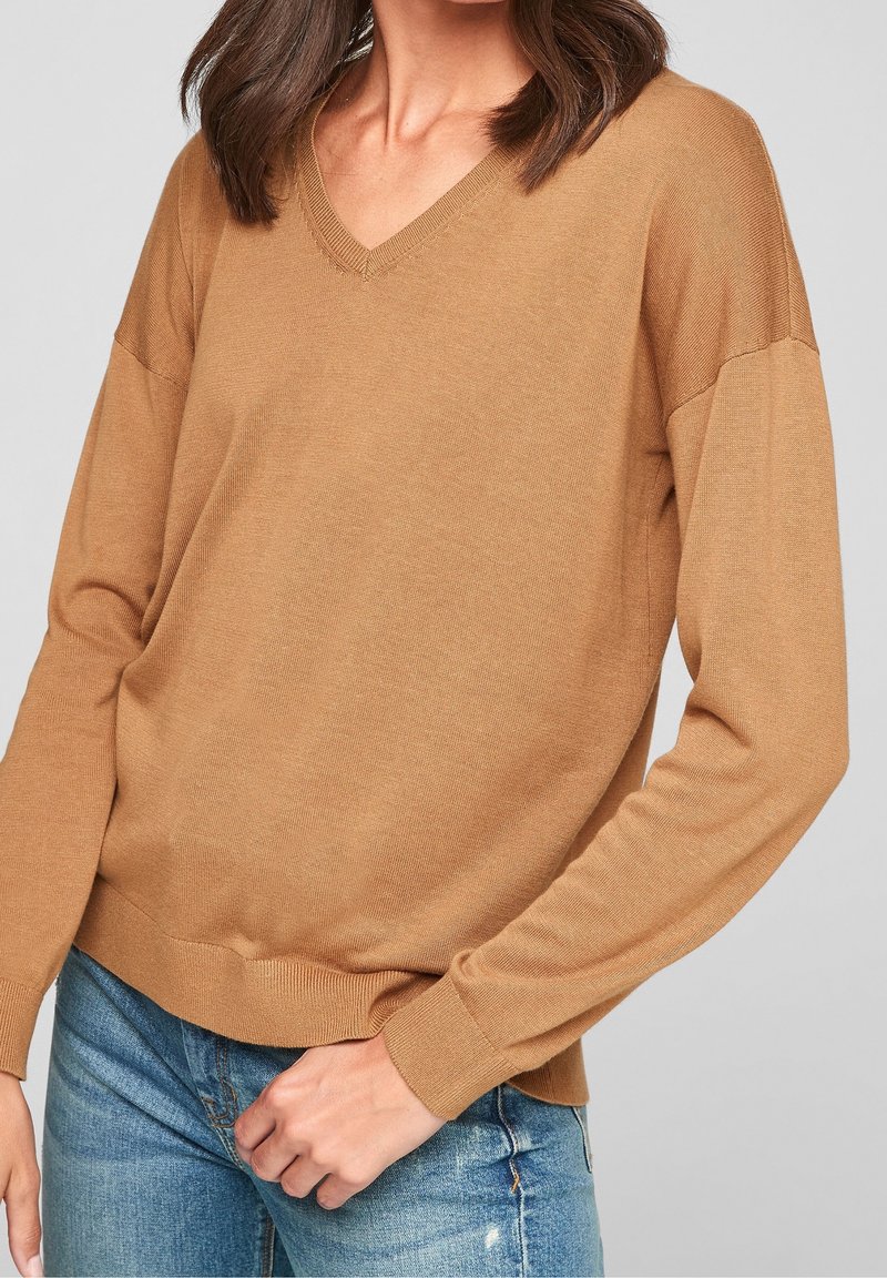 Femme portant un pull col en V couleur chameau à manches longues et un jean bleu, debout devant un fond gris uni.
