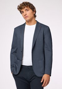 Granatowy żakiet o fakturze z dwoma przednimi kieszeniami, jednorzędowy krój i kontrastujące czerwone przeszycia na klapie, połączony z białą koszulą.
