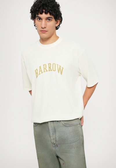 T-shirt a maniche corte color crema con la scritta "BARROW" in lettere dorate, abbinato a jeans leggeri verde chiaro con effetto usurato. Fit casual, texture morbida.