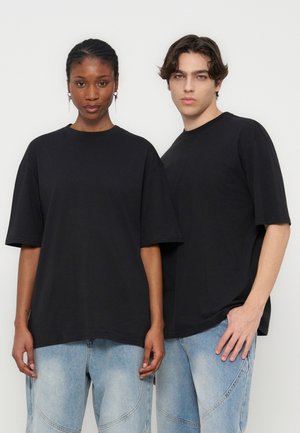 Twee modellen dragen losse zwarte t-shirts en lichtblauwe jeans en staan tegen een effen witte achtergrond, naar voren kijkend.