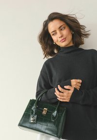Green, croc-embossed handbag with gold hardware and dual handles. Features a structured design and glossy finish. Worn with a black turtleneck sweater.
