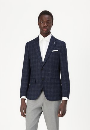 Mørkeblå ternet blazer i et tekstureret stof, med to forlommer, et hvidt lommetørklæde og sorte knapper over en hvid skjorte.