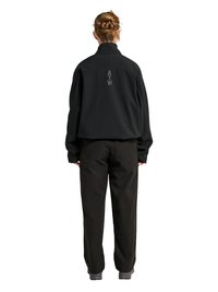 Person mit geflochtenem Haar, von hinten gesehen, trägt eine schwarze kurze Jacke mit dem Schriftzug „ATW“ auf dem Rücken und weite schwarze Hosen.