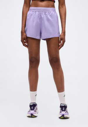 Personne portant un short de sport violet clair, des chaussettes blanches et des chaussures de course violettes, debout avec les jambes droites sur un fond uni.