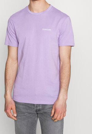 Homme portant un t-shirt à manches courtes lavande avec le texte "YOURTURN" sur la poitrine et un jean gris, debout devant un fond clair uni.