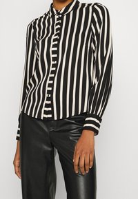 Blusa a righe bianche e nere con colletto, patta bottoni e maniche lunghe. Il tessuto presenta una texture liscia.