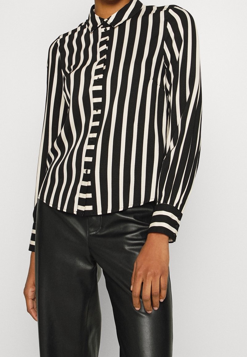 Blusa a righe bianche e nere con colletto, patta bottoni e maniche lunghe. Il tessuto presenta una texture liscia.