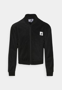 Chaqueta bomber negra hecha de tela suave, con cremallera frontal, puños acanalados y parche con logo en el pecho. Textura lisa y diseño ajustado.