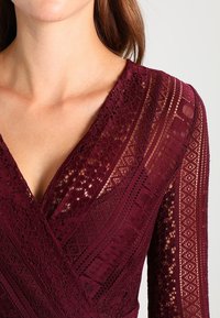 Burgundy spetsklänning med V-ringning, långa ärmar och intrikata mönster med texturerade accenter över tyget.