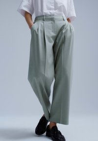 Persoon draagt lichtgrijze wijde broek, wit kortärmelig overhemd ingestopt, zwarte loafers, handen in de zakken, staand tegen een neutrale achtergrond.