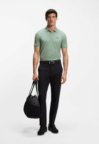 Camiseta polo verde con estampado y mangas cortas, cinturón negro, pantalones negros y zapatillas negras. Sosteniendo una bolsa de deporte negra.