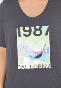 Šedé tričko s V-výstřihem, které obsahuje barevnou grafiku palem, houpací sítě a nápis "1987 CALIFORNIA" tučnými černými písmeny.