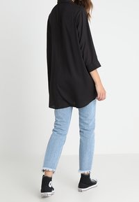 Camicia lunga nera, morbida, con maniche arrotolate, abbinata a jeans azzurri a sigaretta e sneaker neri alti. Design semplice e informale.