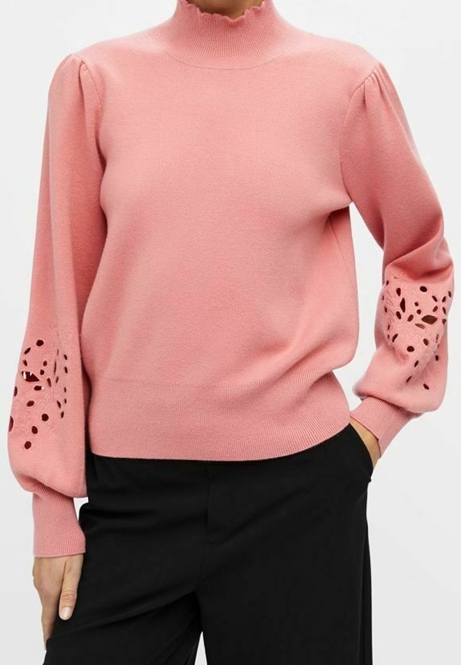 Femme portant un pull rose avec des manches bouffantes présentant des motifs floraux découpés, assorti à un pantalon noir.