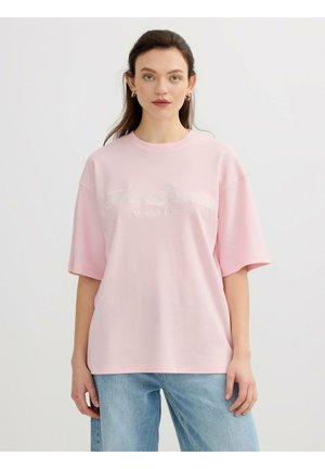 Joven mujer con cabello largo y oscuro que lleva pendientes de aro dorados, una camiseta rosa claro extragrande y vaqueros azules, de pie contra un fondo liso.