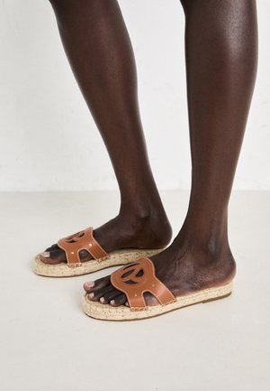 Smeđe kožne natikače s izrezbarenim dizajnom i džute potplate, s zaobljenim vrhom i detaljima šivanja. Potplat ima prirodnu teksturu.