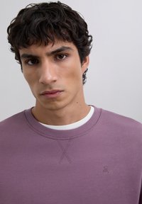 Joven con cabello oscuro y rizado, que lleva un sudadera morada sobre una camisa blanca, contra un fondo claro y sencillo.