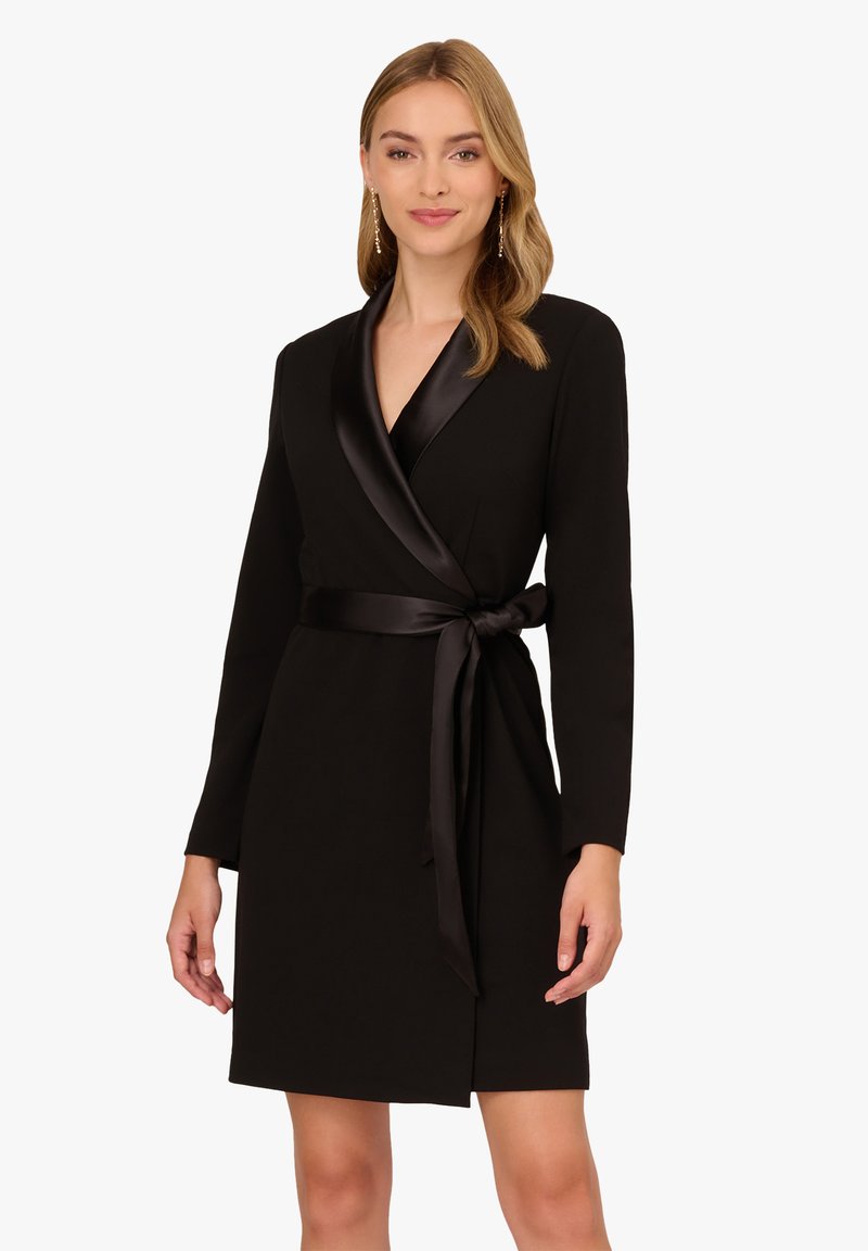 Robe noire à manches longues avec un col châle en satin. Dotée d'une ceinture en tissu à la taille et d'une fente avant subtile.