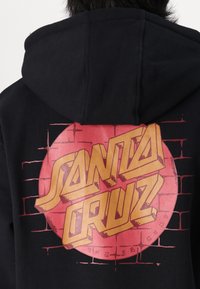 Črn kapuča z velikim rdečim krožnim grafikom na hrbtu in zlatim napisom "SANTA CRUZ" ter oblikami opečnega zidu.