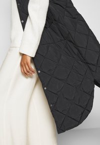 Gepolsterte schwarze Steppweste mit silbernem Zubehör; über einem cremfarbenen Pullover getragen, der einen strukturierten Strickbund und glattes Material darunter zeigt.