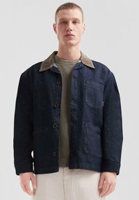 Noor mees kannab tumedat denim-naha jopet, millel on pruun corduroy krae, oliivroheline kudum särgi all ja helesinised püksid, seistes lihtsa tausta against.
