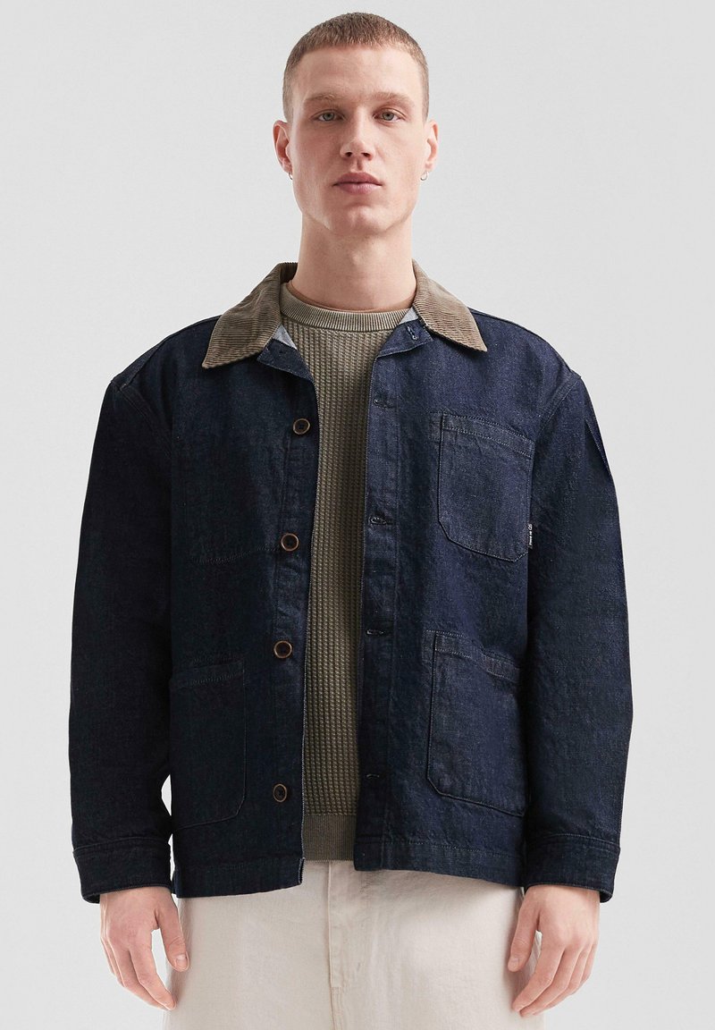 Noor mees kannab tumedat denim-naha jopet, millel on pruun corduroy krae, oliivroheline kudum särgi all ja helesinised püksid, seistes lihtsa tausta against.