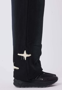 Pantalones negros con bordado de estrellas blancas deshilachadas en el dobladillo, combinados con zapatillas deportivas negras con suela texturizada y acentos en contraste.