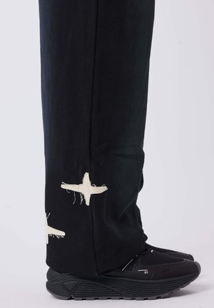 Pantaloni neri con ricamo di stelle bianche sfrangiate all'orlo, abbinati a scarpe sportive nere con suola testurizzata e accenti a contrasto.