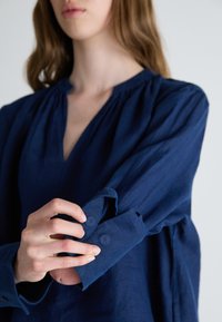 Camicia di lino blu navy con scollo a V e dettagli arricciati. Le maniche lunghe hanno polsini con bottoni. Tessuto morbido visibile nel materiale.