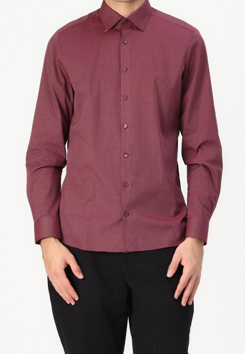 Homme portant une chemise habillée bordeaux à manches longues, boutonnée, avec un col pointu et un pantalon noir, debout, les bras détendus.
