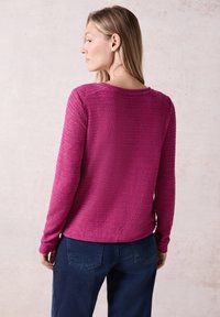 Maglietta a maniche lunghe in tessuto magenta texturizzato con scollatura rotonda e righe orizzontali; abbinata a pantaloni blu scuro, vista da dietro.