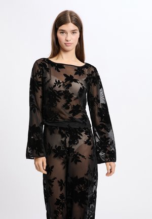 Top de superposición negra transparente con patrón floral, mangas largas y un escote redondo, que presenta una textura suave y un delicado detalle.